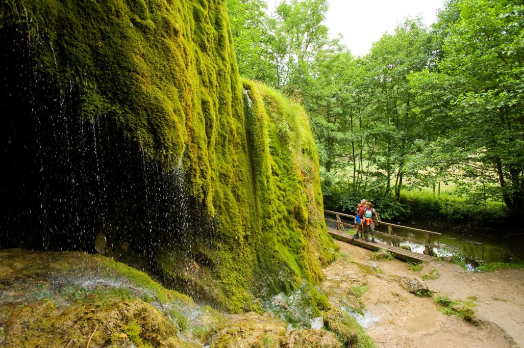Wasserfall mit moosbedecktem Felsen und Person am Ufer eines Bachs.