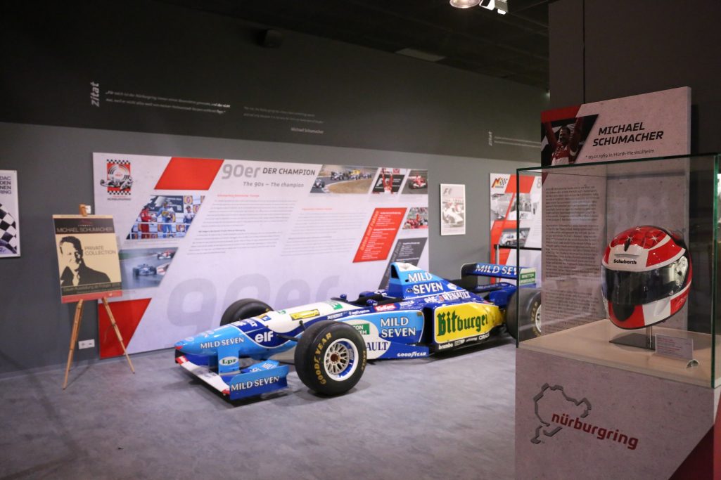 Ein blauer Rennwagen steht in einem Ausstellungsraum neben einem Helmdisplay.