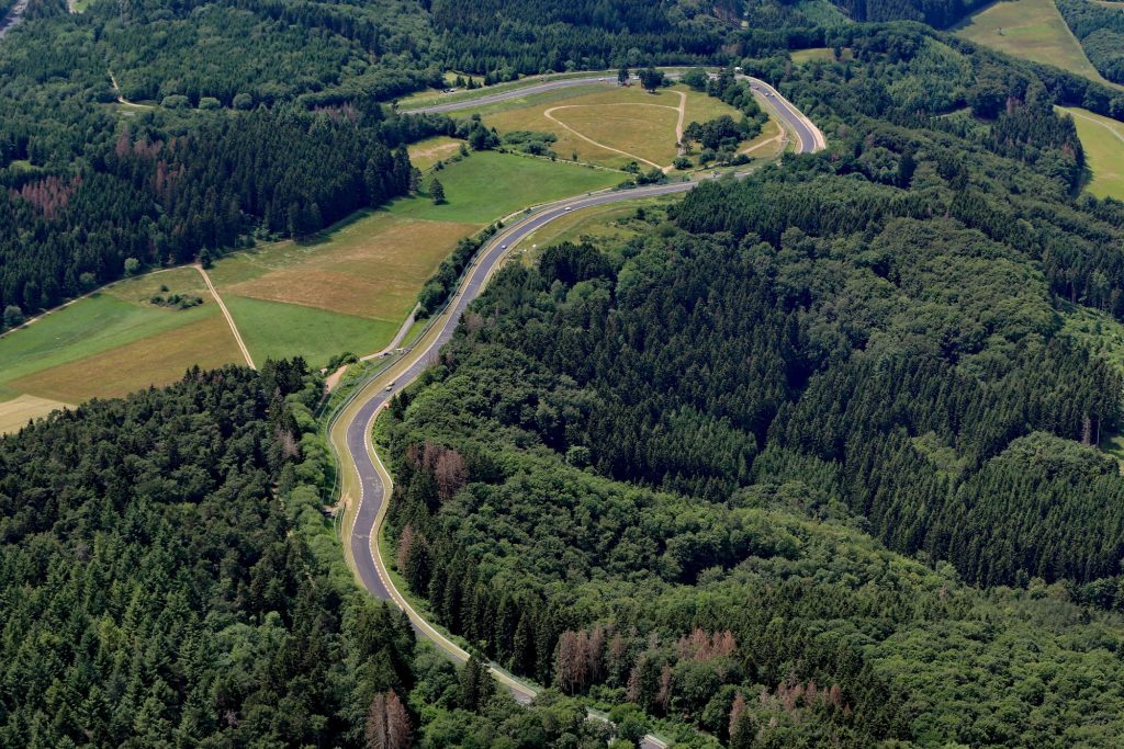 Kurvige Straße durch bewaldete Landschaft mit Wiesen und Feldern im Hintergrund.