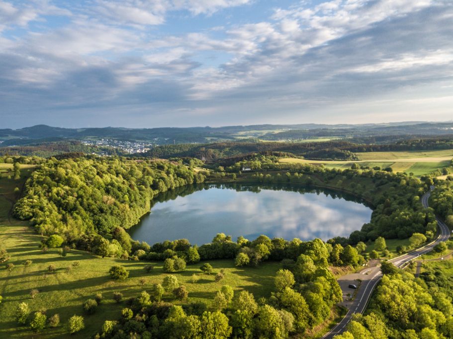 Landschaft mit einem runden See, umgeben von Bäumen und Wiesen unter einem bewölkten Himmel.